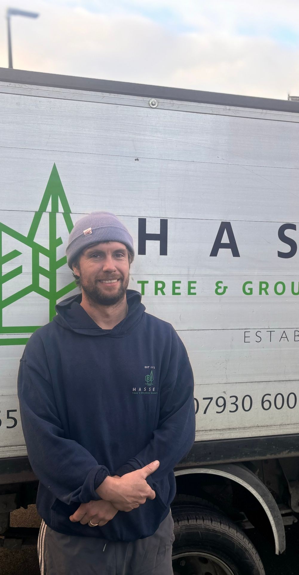 Person wearing a beanie and dark hoodie standing in front of a truck with business signage.